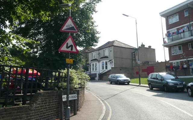 Warning signs on Picardy Road in Belvedere for a 10% steep hill and a double bend, with the road curving away past parked cars